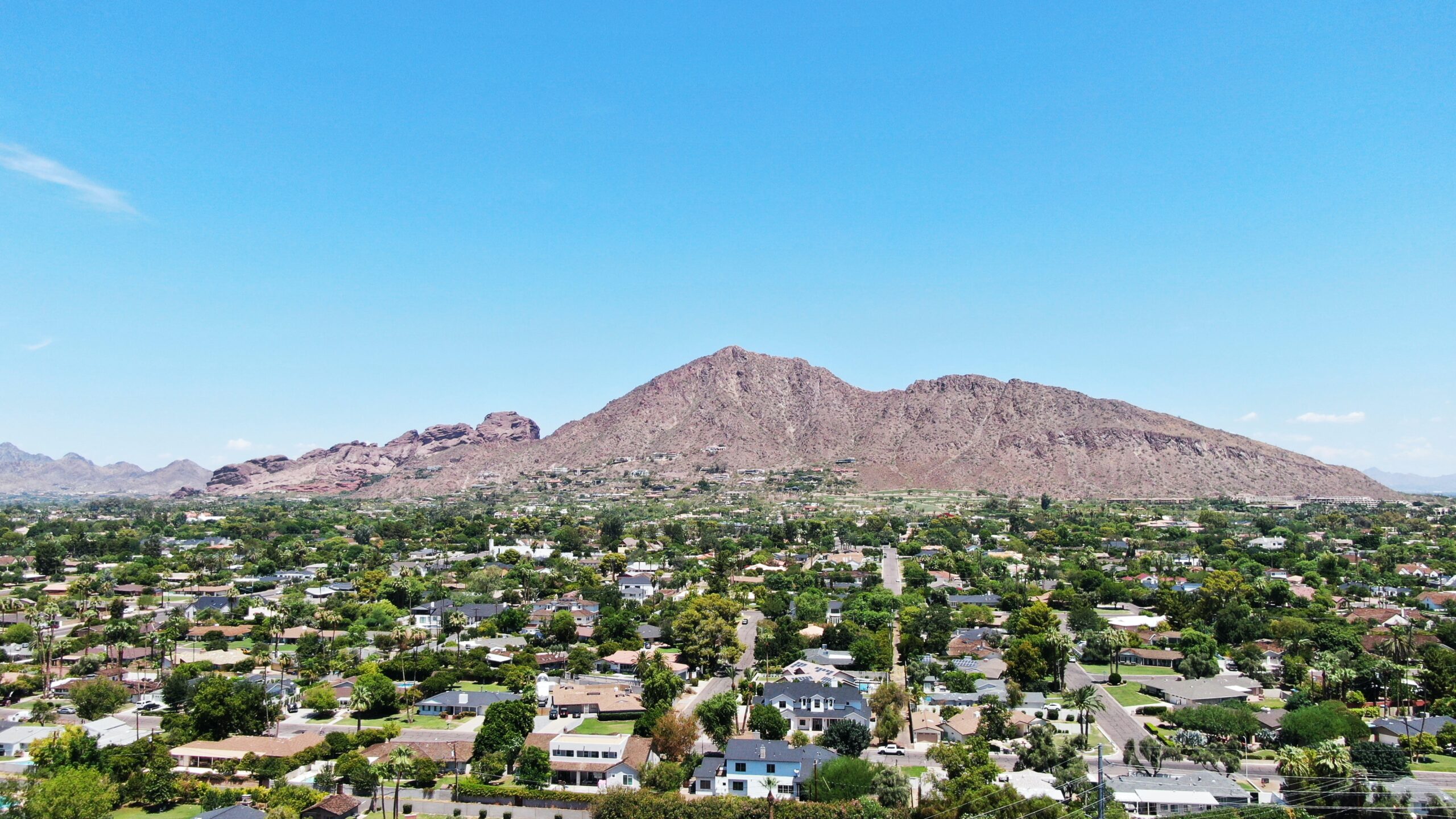 Aerial overview of a tree-filled residential area in Camelback Mountain located in Phoeniz Arizon. There are clear blue skies and mountains in the back, overlooking the neighborhood.