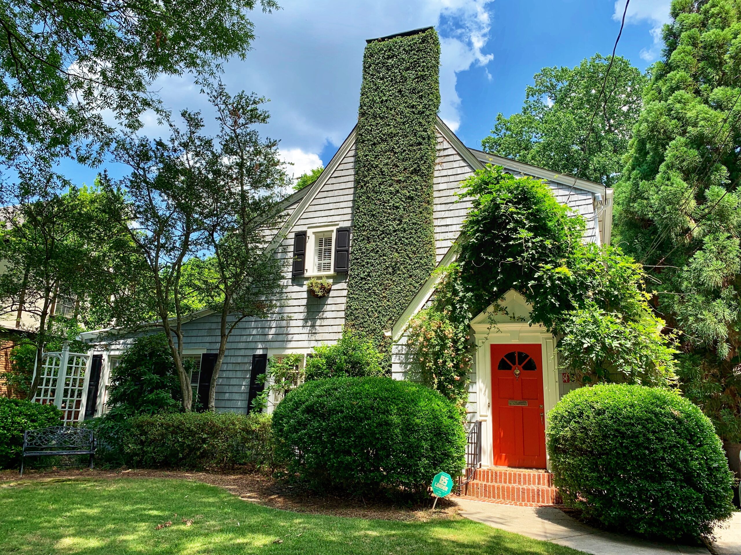 White home with red door surrounded by lush greenery, trees, and sculpted bushes in the summertime