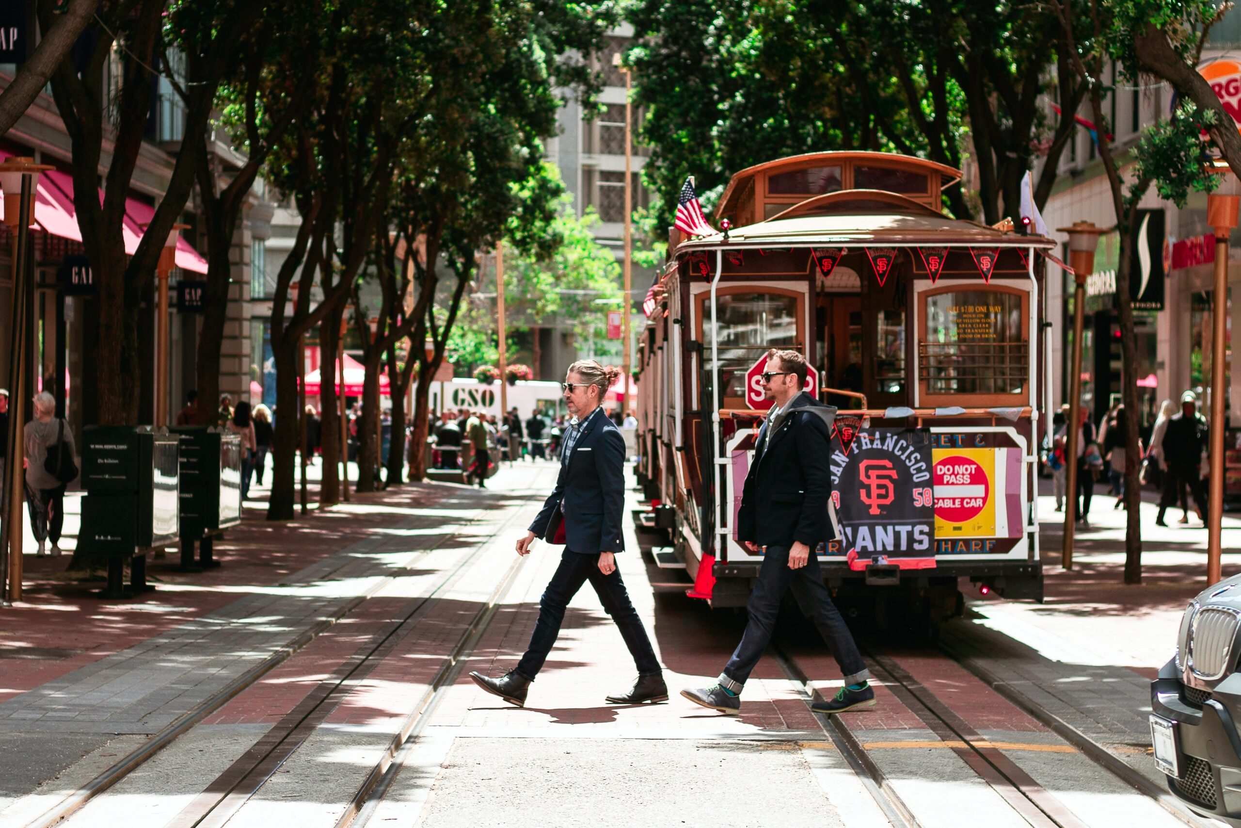 Pedestrian-friendly area with people walking to cross a tree-lined street in front of a street car on a bright, sunny day